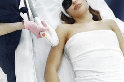Crop faceless beautician conducting laser hair removal to young lady in safety glasses in clinic